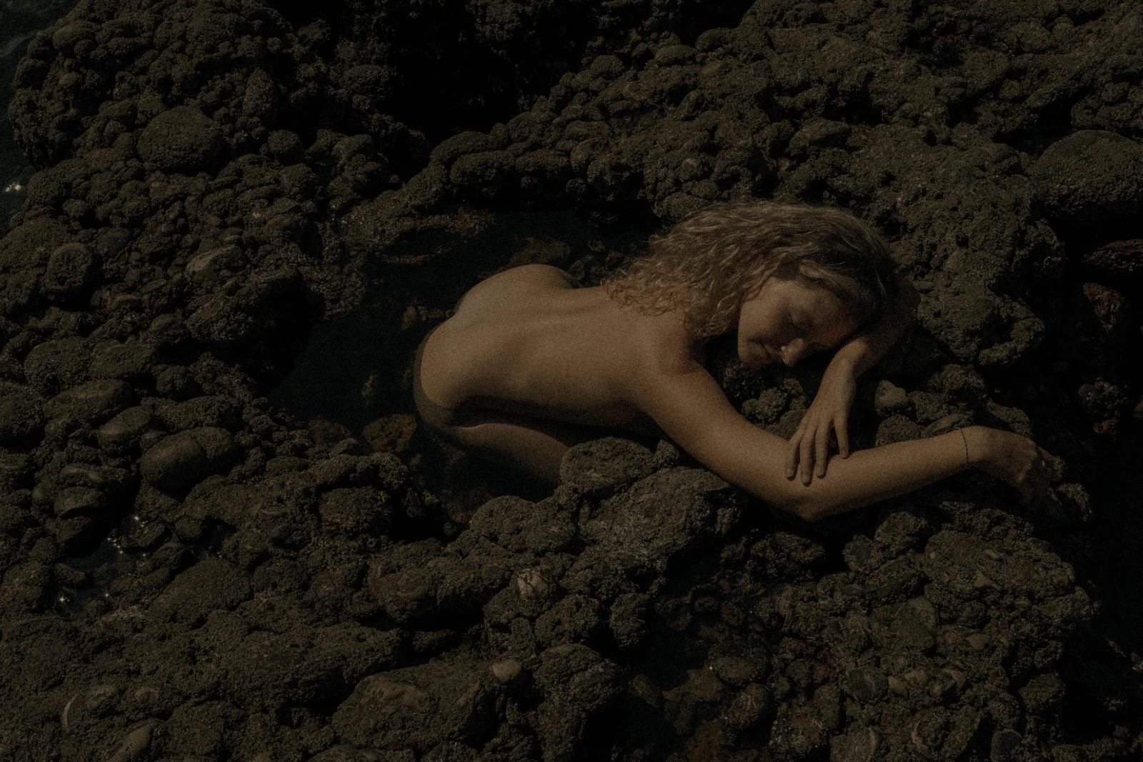 A figure rests with their head on one arm among dark coastal rocks.